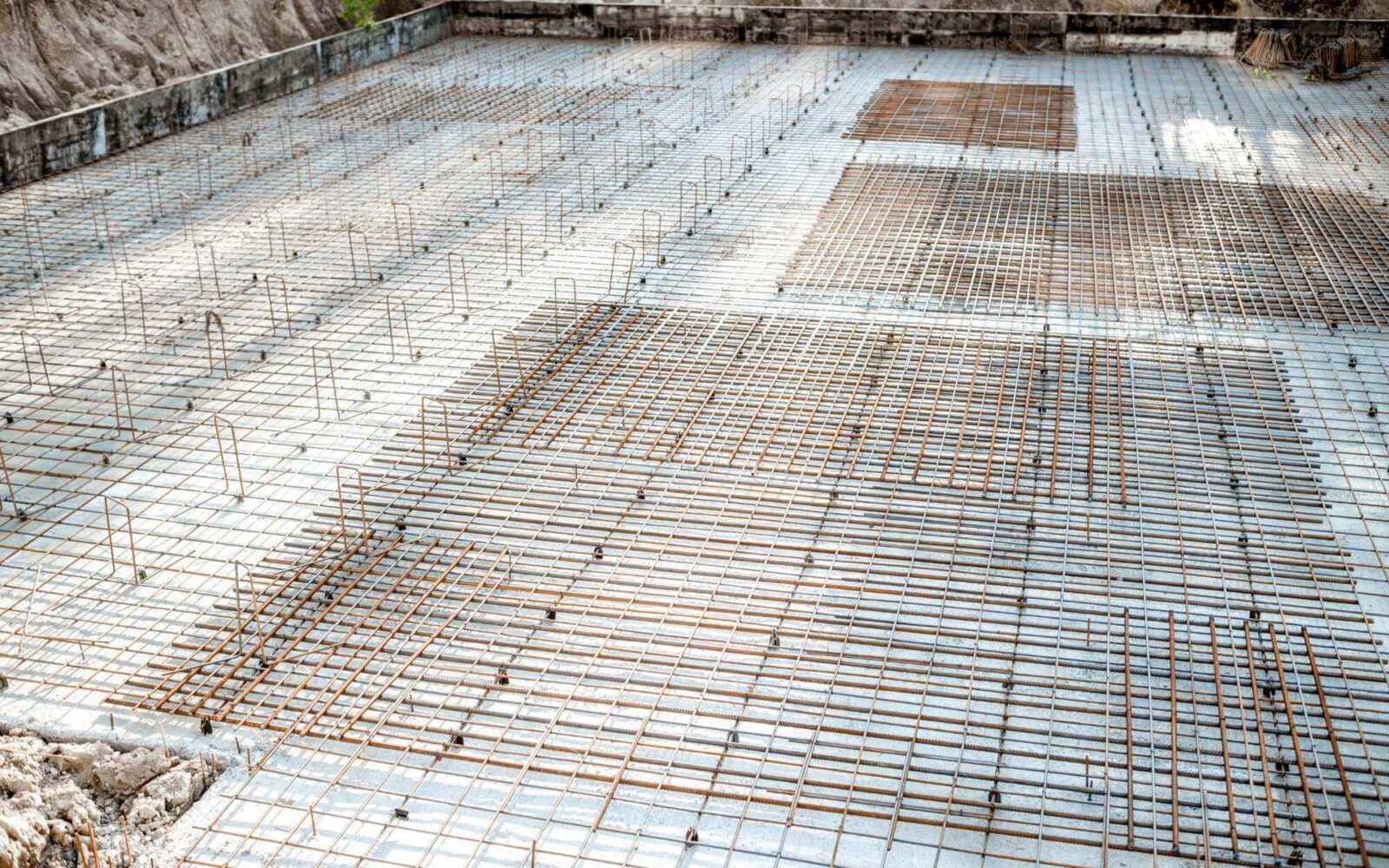 The image shows a rectangular construction site foundation in Phoenix, AZ, covered with a grid of rebar (reinforcing steel bars) in a symmetrical pattern. The groundwork, meticulously prepared by a foundation contractor, is ready for pouring the concrete slab, with visible rebar and spacers maintaining an even layout.
