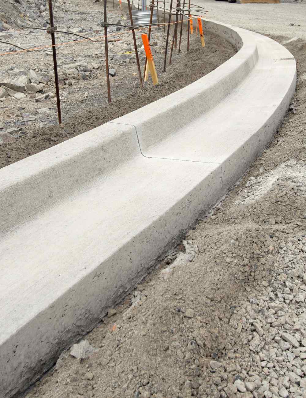A newly poured concrete curb curves sharply on a construction site in Gilbert, AZ. The area around the curb is unpaved, with dirt and gravel. Wooden stakes with safety flags are positioned along the curb, and rebar sticks out of the ground nearby, revealing the meticulous work of East Valley Concrete.
