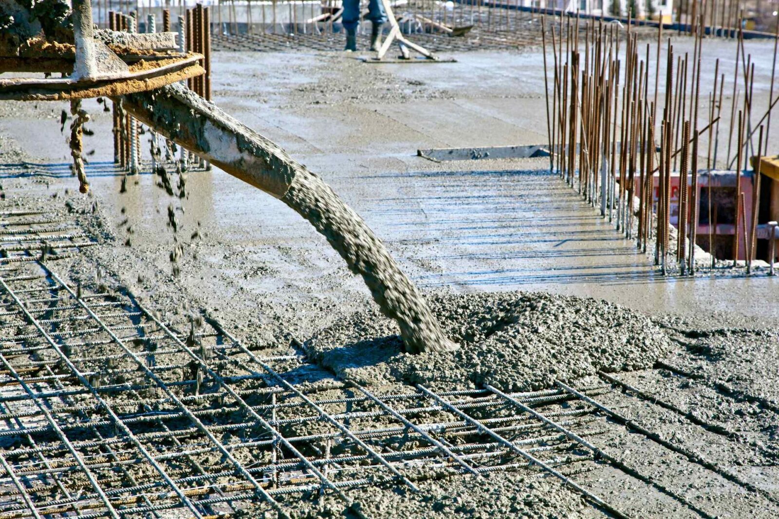 A concrete contractor in Gilbert AZ operates a pump, discharging wet concrete onto the construction site and filling a steel rebar framework. Workers in the background attend to leveling and smoothing the freshly poured concrete. The scene shows a work in progress on a structural slab. Free quotes available!