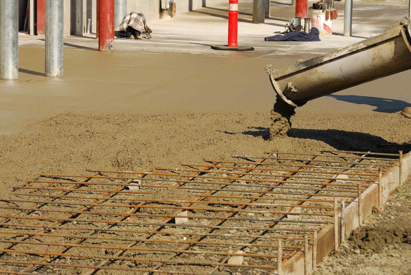 Concrete is being poured from a chute onto a construction site in Mesa, AZ, covering a grid of steel rebar. The surrounding area shows construction materials and equipment, managed by licensed concrete contractors.