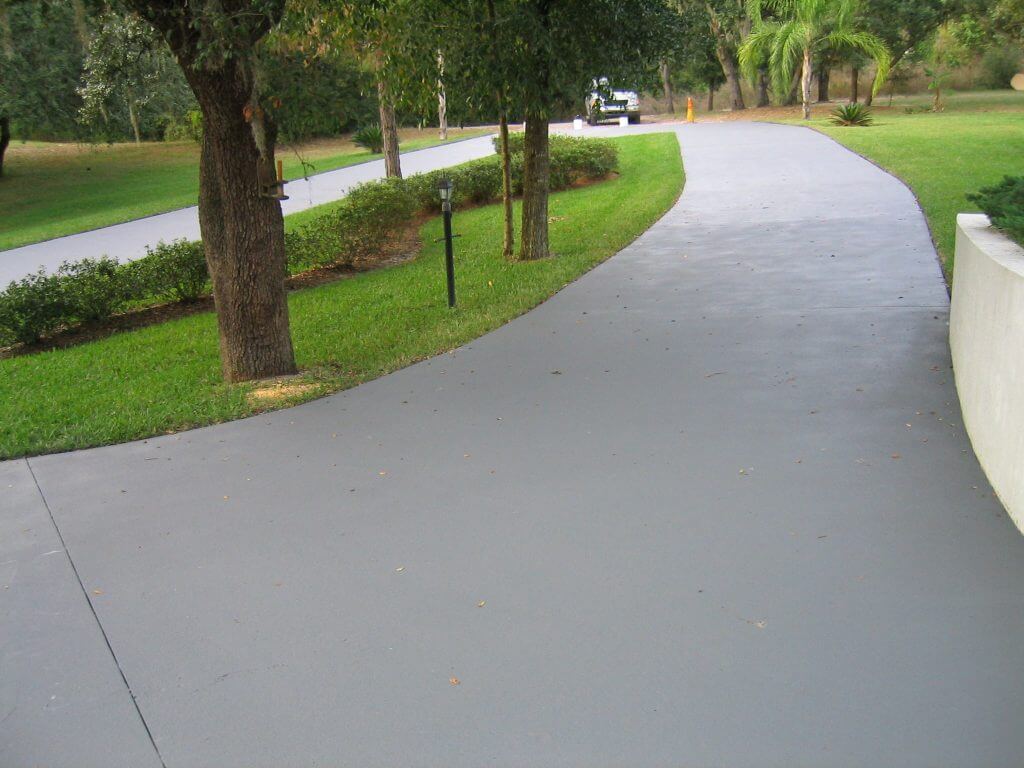 A smooth, wide concrete pathway winds through a well-maintained garden with lush green grass and trees. A golf cart is visible in the distance, parked on the path. Upgrading to durable surfaces here ensures lasting beauty and convenience. A white curved wall borders part of the path on the right side.