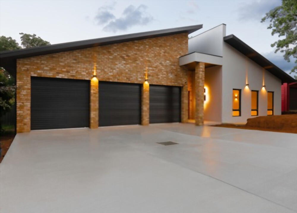 A modern house with a sloped roof features a blend of brick and white panel exteriors, constructed by the renowned Chandler AZ concrete contractor, East Valley Concrete. The front facade has three black garage doors, each illuminated by wall-mounted lights. Several narrow windows are adjacent to a recessed entryway with a small overhang.