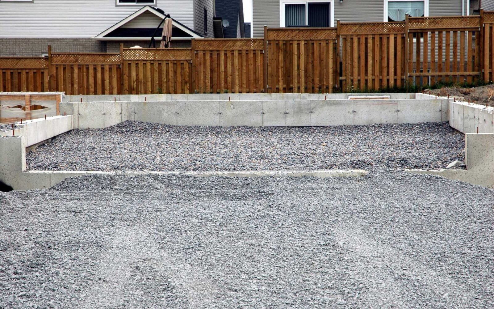 A concrete slab for a new house is surrounded by gravel and forms the base of a construction site in Phoenix AZ. In the background, there are wooden fences and neighboring homes. The foundation contractor has prepared the site for further building work.