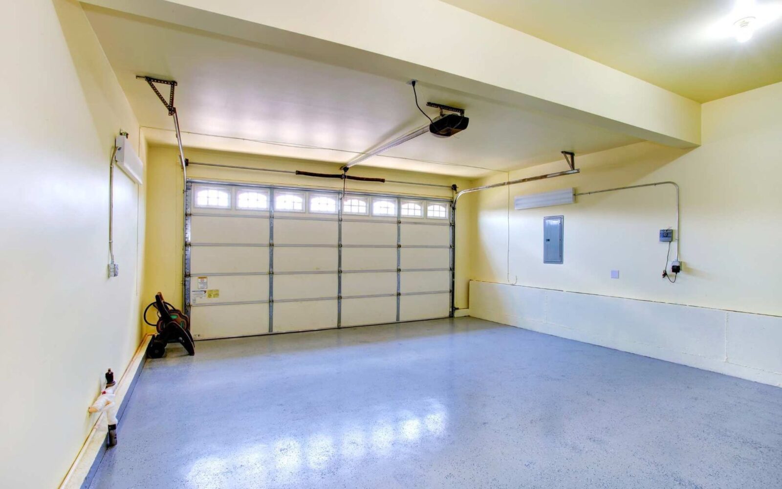 A clean, empty garage with a polished light blue concrete slab floor and cream-colored walls. There is a large closed garage door with rectangular windows at the top. The garage has a ceiling-mounted opener, a wall-mounted vacuum cleaner, and visible wiring and outlets.