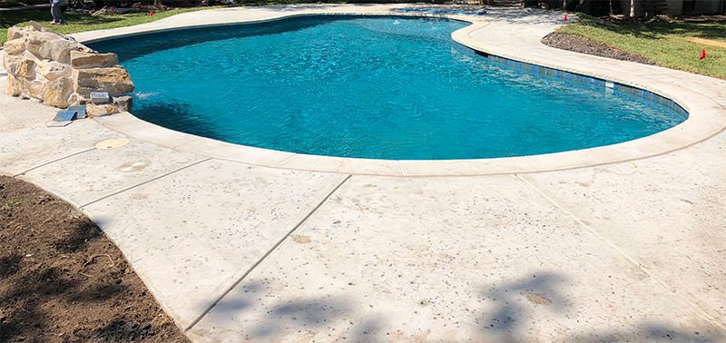 A serene backyard pool with clear, blue water is surrounded by a light-colored concrete deck crafted by East Valley Concrete, a trusted concrete contractor in Gilbert, AZ. Small, decorative rocks adorn one side of the pool. The grassy area around it has some patches of dirt, and shadows of nearby trees are cast on the deck.