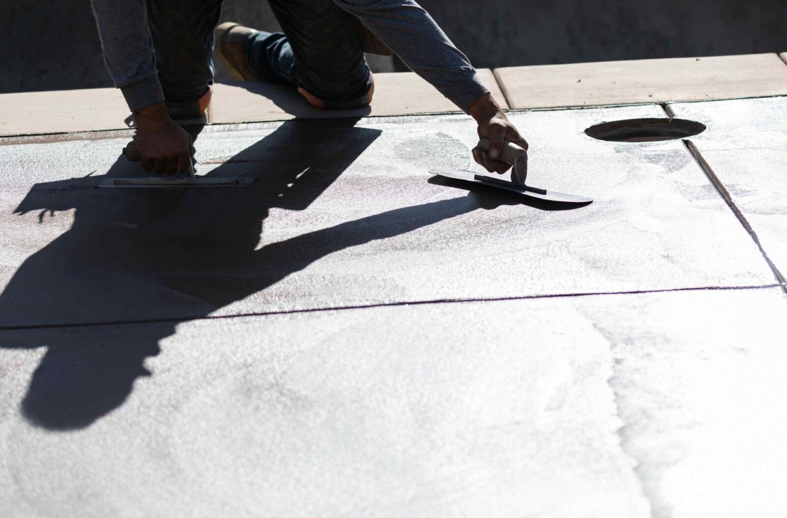 A person kneels on a concrete surface while using trowels to smooth the concrete. The angle creates a stark shadow of their hands and tools on the ground. The background is slightly blurred, with an opening on the right side—work typical of East Valley Concrete, your trusted concrete contractor offering free quotes.