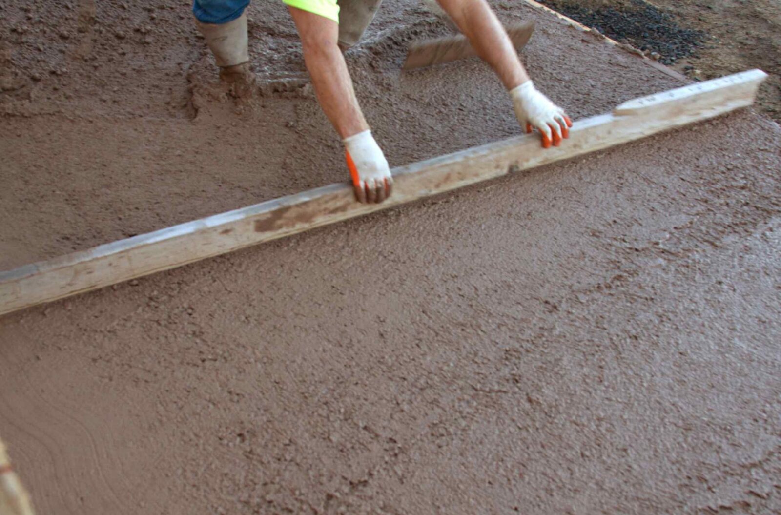 A person wearing a yellow shirt and gloves levels wet concrete using a large wooden plank. The image captures the person's arms and the surface of the freshly poured concrete as it is being smoothed by East Valley Concrete to meet Tempe AZ's concrete needs.
