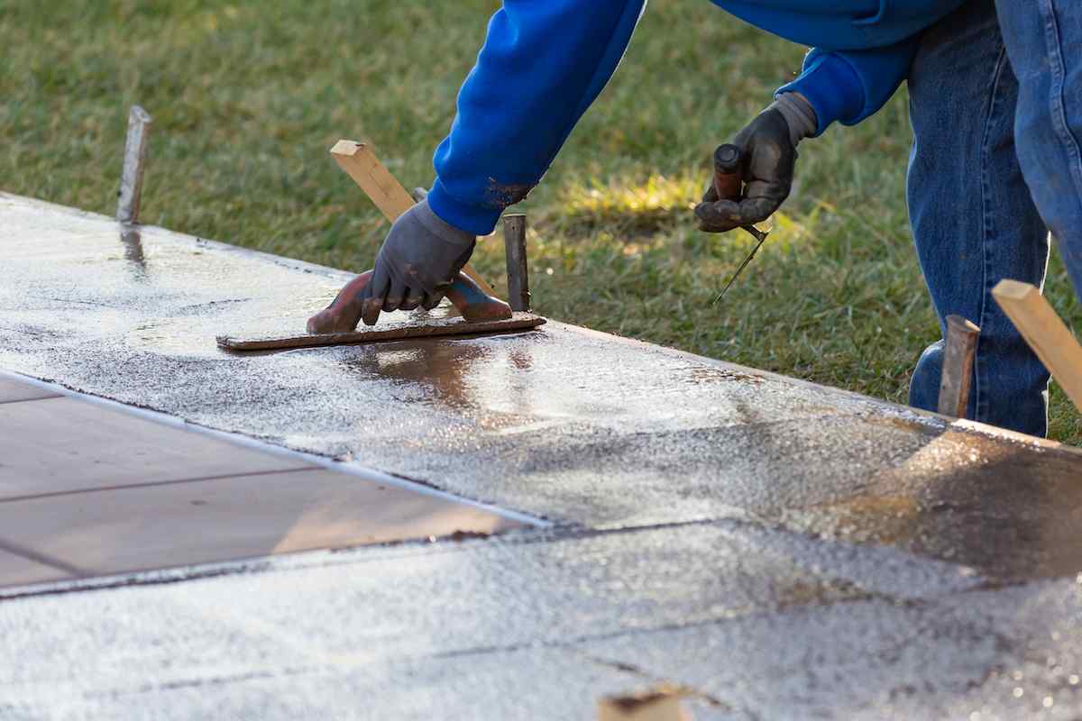 A person wearing a blue hoodie and gloves smooths wet concrete on a surface, using a trowel. The individual is working outdoors on a grass lawn in Tempe AZ, with wooden stakes visible around the concrete area. The project appears to be managed by East Valley Concrete, known for their precise work in the region.