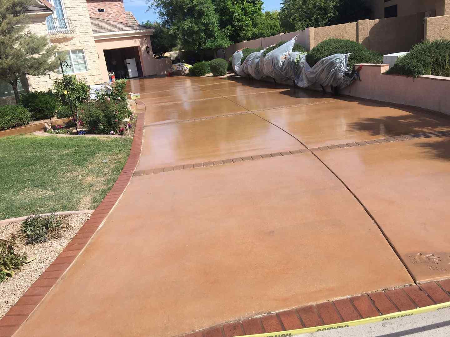 A wide, freshly-paved driveway by East Valley Concrete leads to a garage of a beige house with brick accents in Chandler, AZ. The driveway is bordered by grass, small shrubs, and a brown brick wall on one side. Covered items and plants line the wall, protected by plastic sheets. Yellow caution tape is visible.