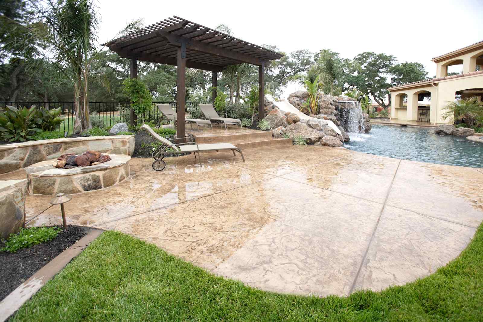 A backyard designed by East Valley Concrete features a stamped concrete patio with a lounge chair, a pergola with two additional lounge chairs beneath it, and a built-in stone fire pit. The patio, crafted by this esteemed pool deck contractor from East Valley Arizona, overlooks a lagoon-style swimming pool with a rock waterfall and lush landscaping surrounding it.