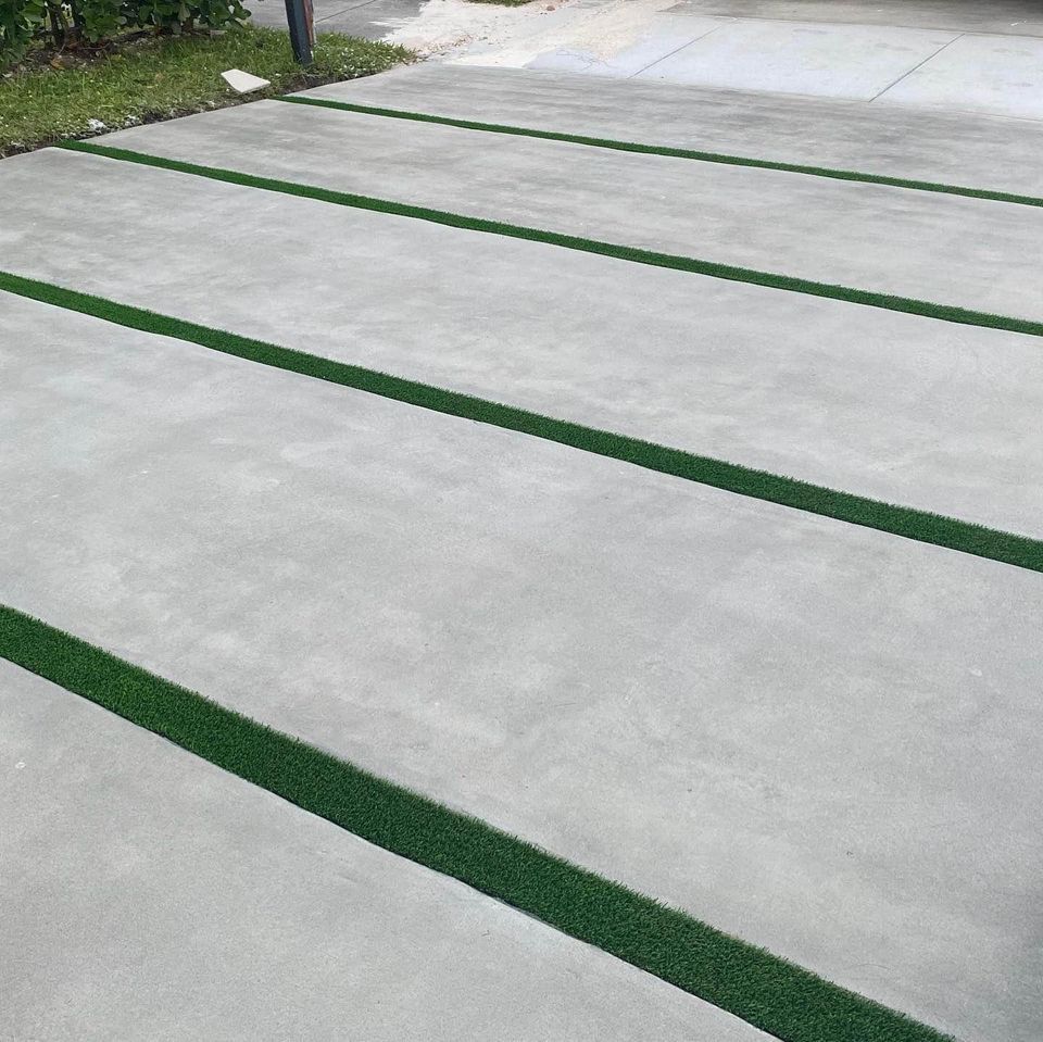 A concrete driveway with wide, flat sections separated by narrow strips of artificial green grass. The design creates a clean, geometric pattern that contrasts the grey concrete with the vibrant green grass. Some foliage and a walkway can be seen in the background. Free quotes available from decorative concrete contractors!