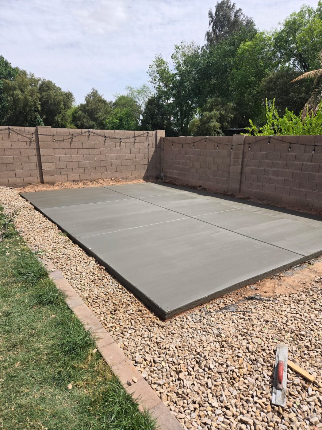 A backyard patio with a reddish-brown concrete surface adjoins a brick house. Crafted by East Valley Concrete, the patio is bordered by curved black trim and a well-maintained garden bed with green plants and shrubs that follow the curve of the patio. This picturesque scene exemplifies quality work in Gilbert, AZ.